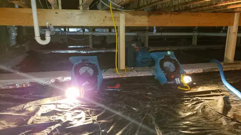 Two air movers drying a crawl space with plastic sheeting on the ground under the floor joists