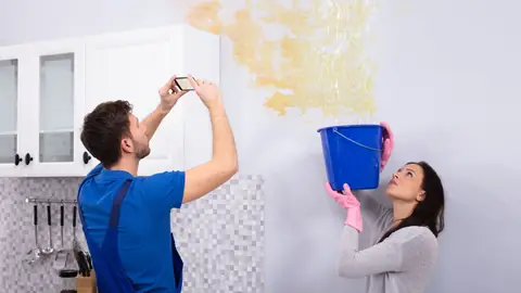 Homeowner photographing a water stain on the ceiling while a bucket catches a leak